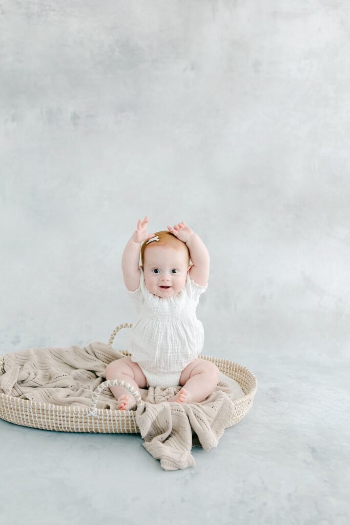 Smiling baby with red hair sitting in a woven basket during a milestone photo session in a Port Saint Lucie studio, wearing a white knit romper against a soft blue backdrop